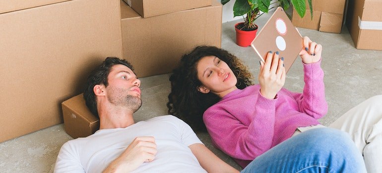 a man and a woman lying on the floor next to cardboard boxes and reading on a tablet