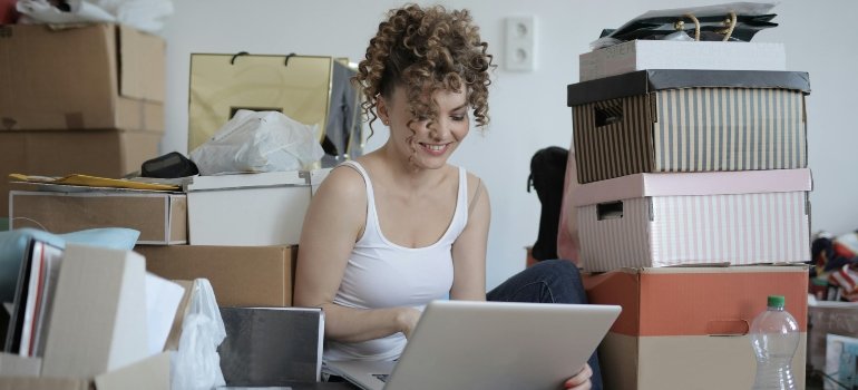 A woman surrounded with cardboard boxes using her laptop while thinking about warning signs that you have too much stuff.