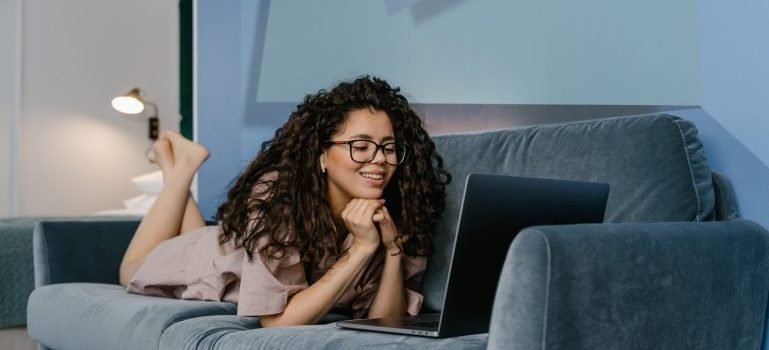 A young woman lying on the couch using her laptop and smiling while reading about moving from Morrisania to Upper East Side.