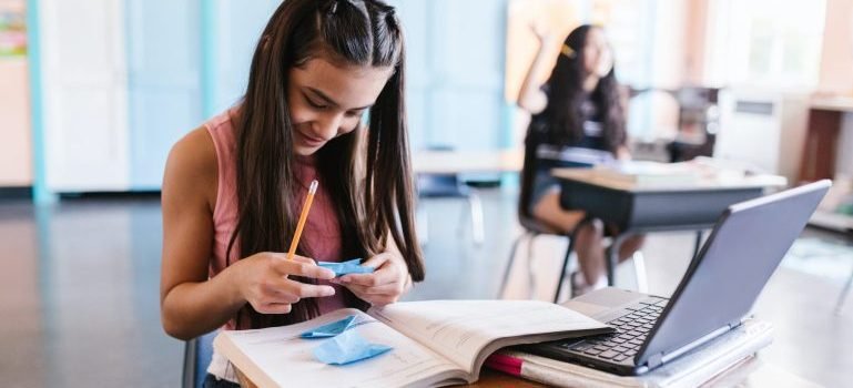 Two girls in the classroom, one in the front holding and reading a from a piece of paper, books and a laptop on the desk.