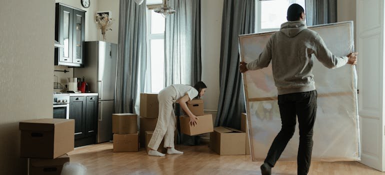 A man holding a big painting and a woman piling some cardboard moving boxes.