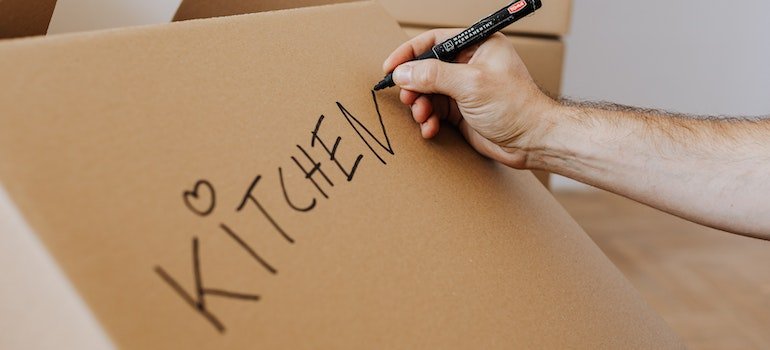 A person writing the word kitchen on a cardboard box.