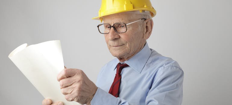 An elderly man with a yellow helmet holding a white piece of paper.