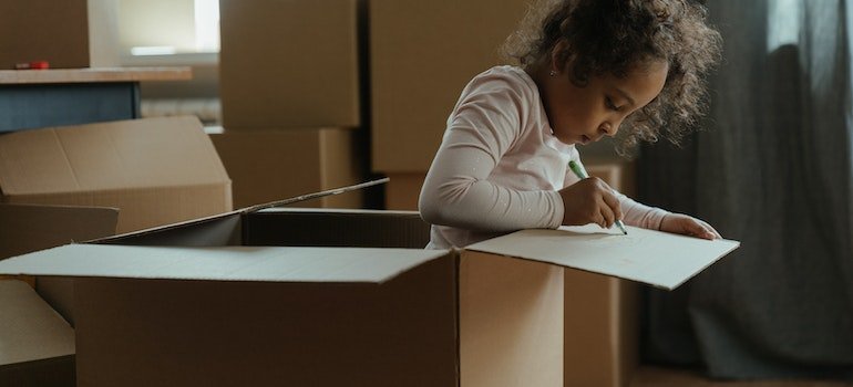 A little girl peaking out of a moving box and drawing something on it.
