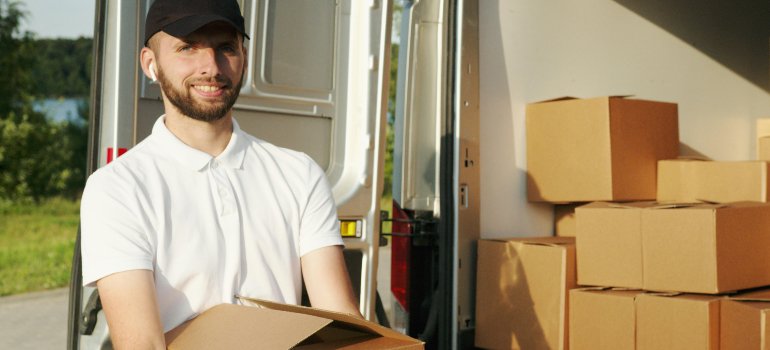 A smiling delivery worker carrying a box next to a van filled with packed boxes.