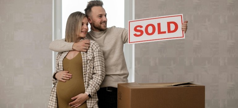 A smiling couple holding a "SOLD" sign in front of moving boxes, excited to start a new chapter in NYC.