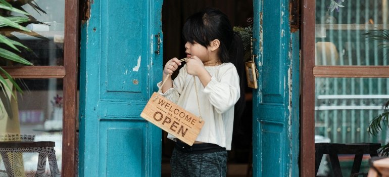 A young girl holding an "Open" sign at the entrance of a small Brooklyn shop.