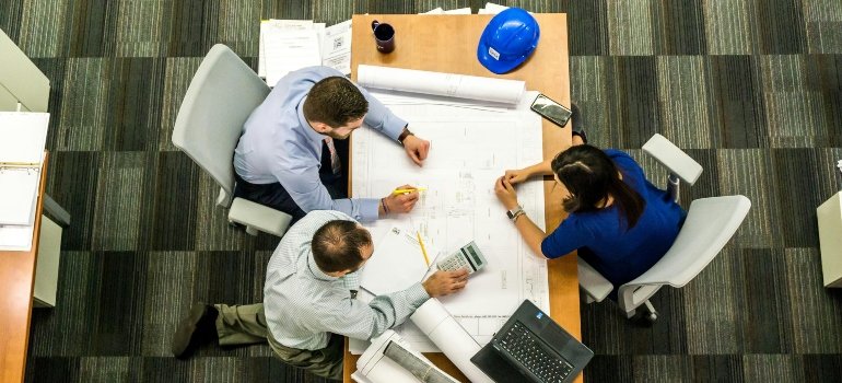 Three colleagues reviewing building plans and brainstorming storage solutions for Cconstruction projects around a desk.