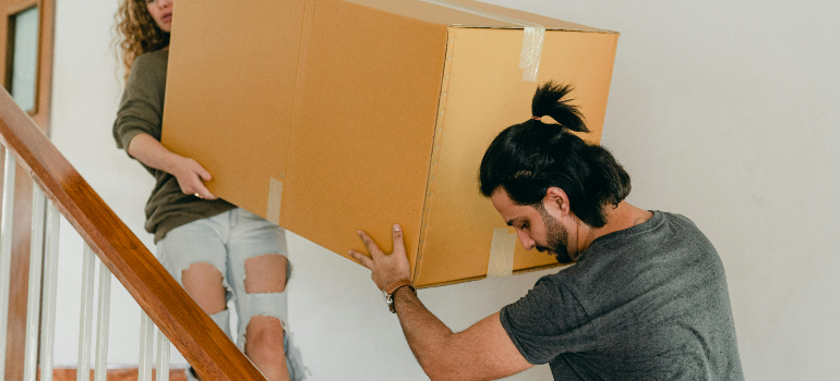 A couple carrying a big cardboard moving box up the stairs.
