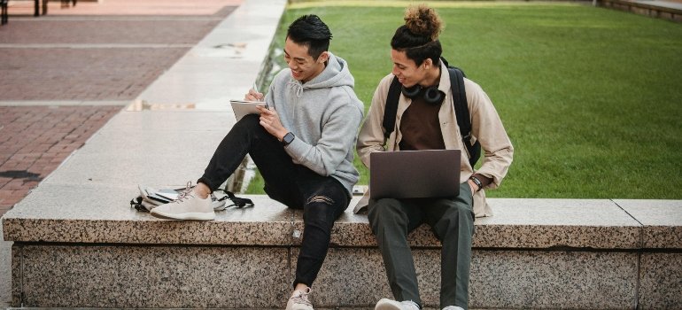 Two male college students collaborating outdoors with laptop and notebook, discussing ideas.