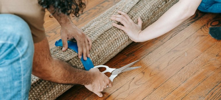 Two people rolling and cutting a rug with scissors on a wooden floor while preparing for a move.