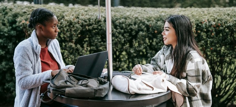 Two female students chatting and studying outside on campus, with backpacks and laptops.