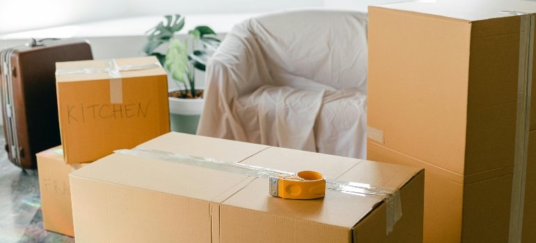 Neatly sealed cardboard boxes and packing tape sit in a bright room with a covered chair, indicating an organized packing process.