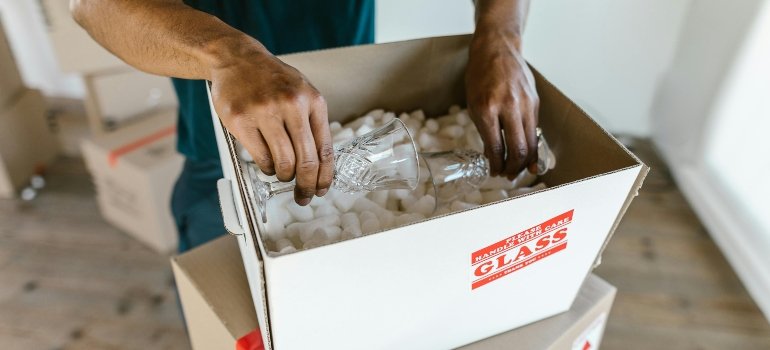 A person carefully placing crystal glasses into a box labeled "fragile" with protective packing peanuts.