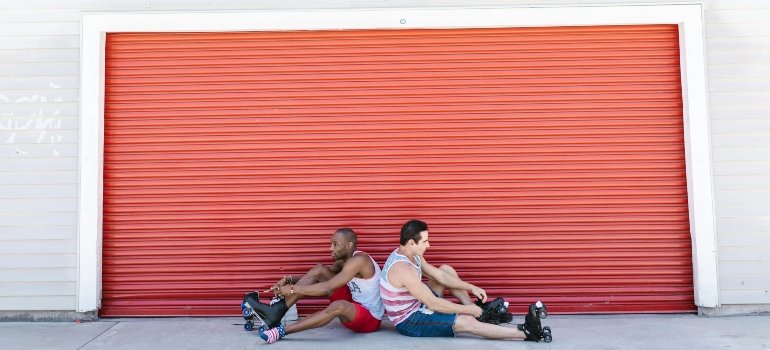 Two men sitting back-to-back on roller skates in front of a bright red garage door, enjoying a sunny moment.