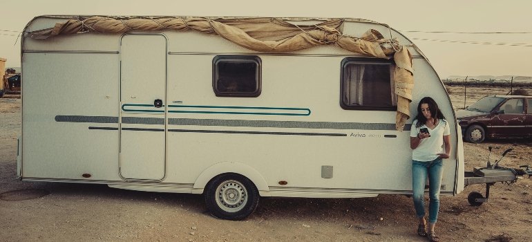 Woman stands beside a white RV parked in a dry, desert-like area, checking her phone with confidence.
