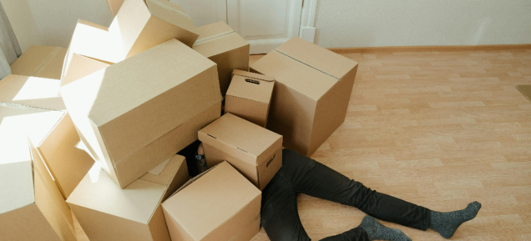 A man lying on the floor under a pile of cardboard moving boxes.