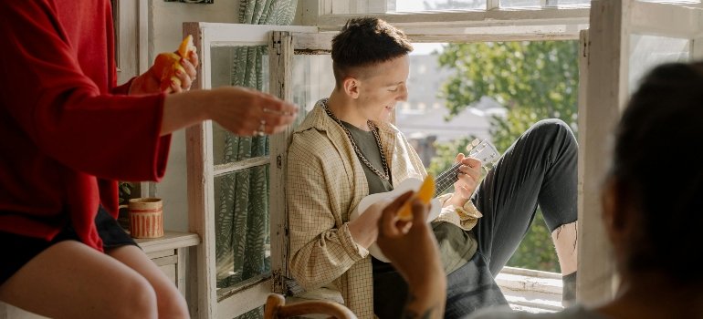 A young man sits by the window playing music while others enjoy snacks in a shared apartment.