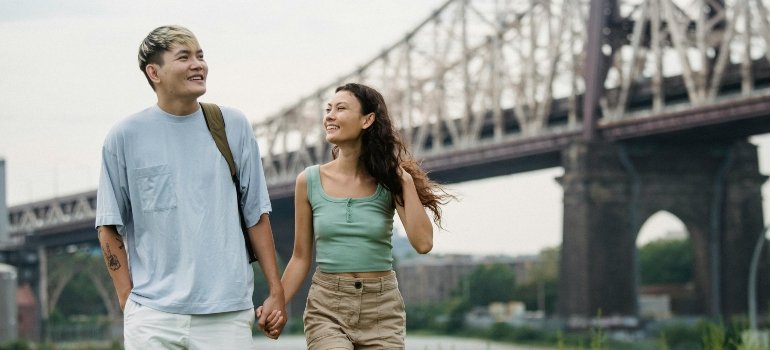 Smiling couple holding hands while walking through a grassy area with a bridge in the background in NYC.