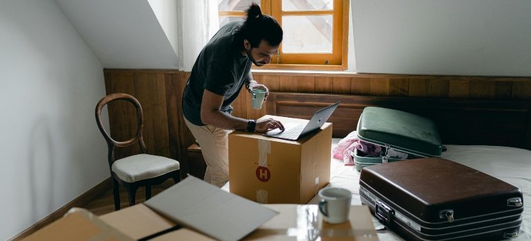 A man balancing coffee and work while surrounded by cardboard chaos.